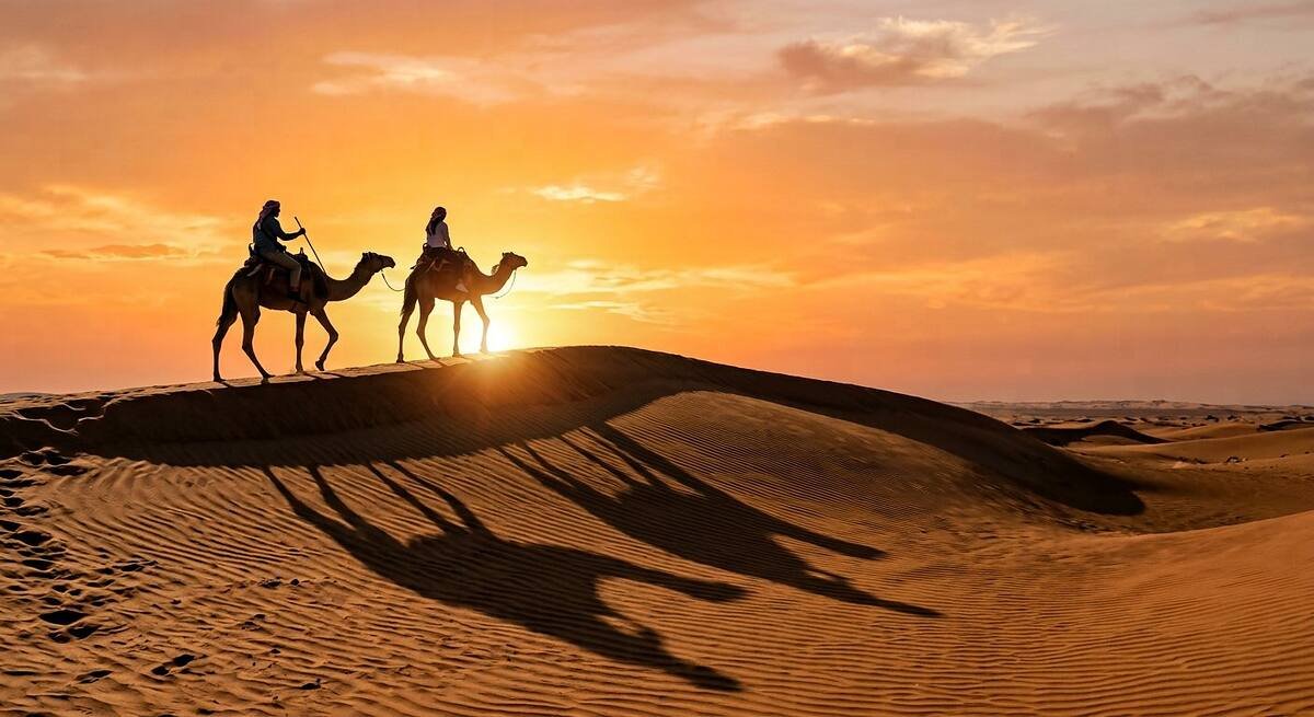 Two guests riding camels along a dune crest at golden hour